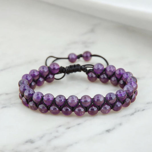 Handmade amethyst beaded macrame bracelet with double row design and adjustable black cord, displayed on a white marble surface.